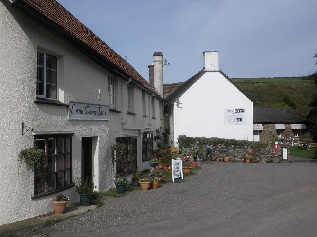 A rural farm shop, 'Lorna Doone Farm', with white buildings, displaying potted plants, a gift shop, and a cafe sign.